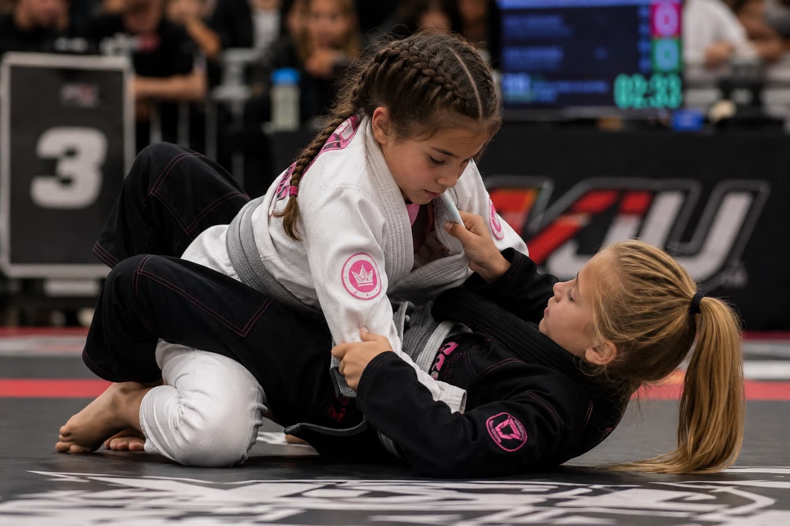 Two young girls in white and black gis grappling in a kids Brazilian Jiu Jitsu tournament match, one working from the top position while the other defends from guard.